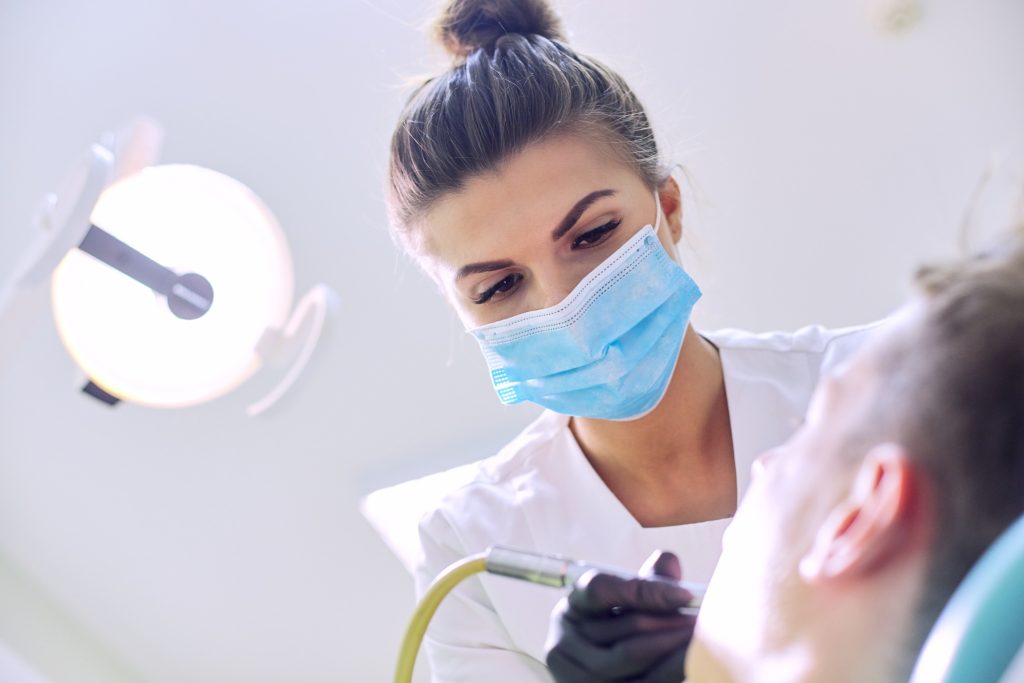 Female Dentist Treating Teeth To Patient, Young Man In Chair At Dental Clinic